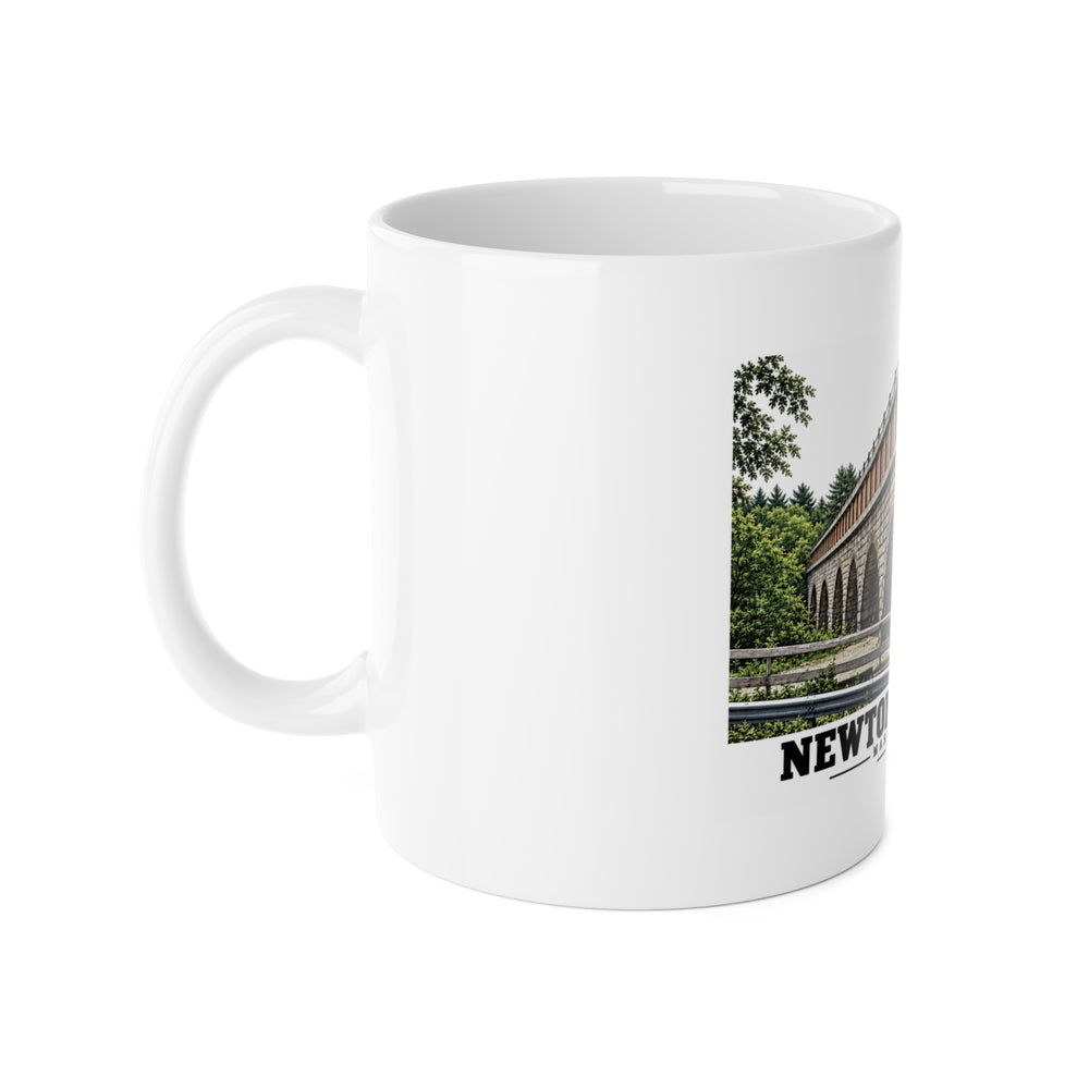 White ceramic mug with its handle on the left and part of the Echo Bridge artwork showing a row of stone arches and a brick upper band, with Newton/Needham lettering visible on the right.