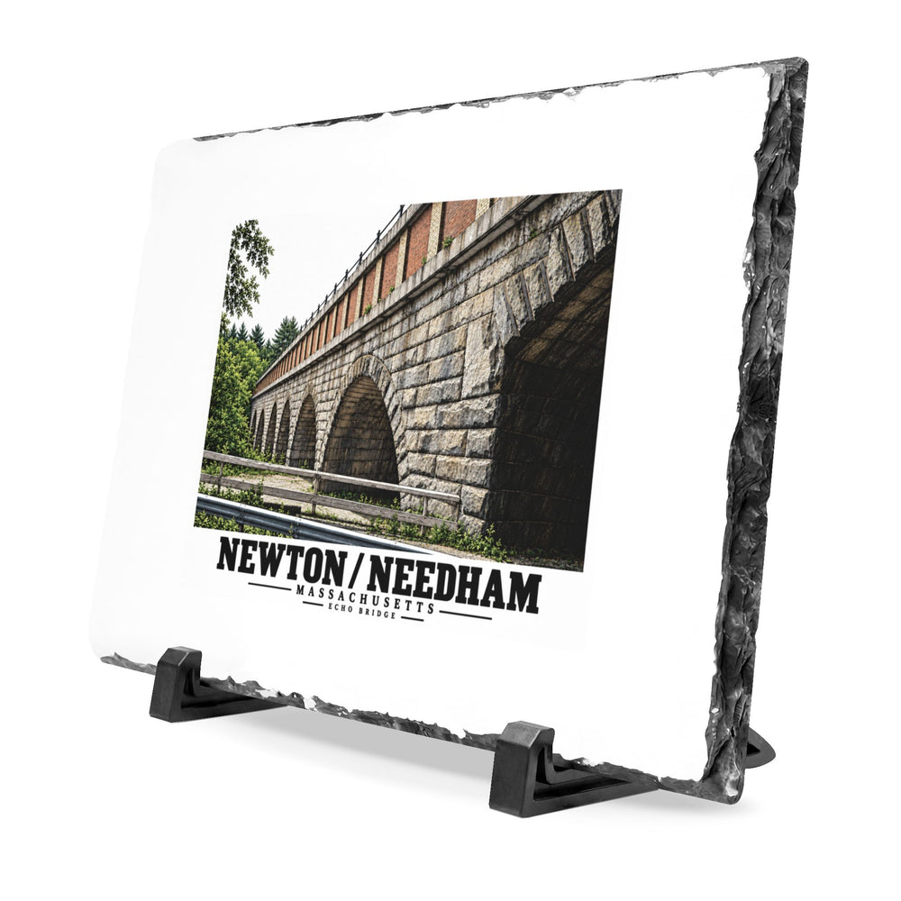 Angled slate plaque on two black supports with Echo Bridge artwork showing a row of stone arches and a brick upper band, with Newton/Needham lettering and a rough dark stone edge.