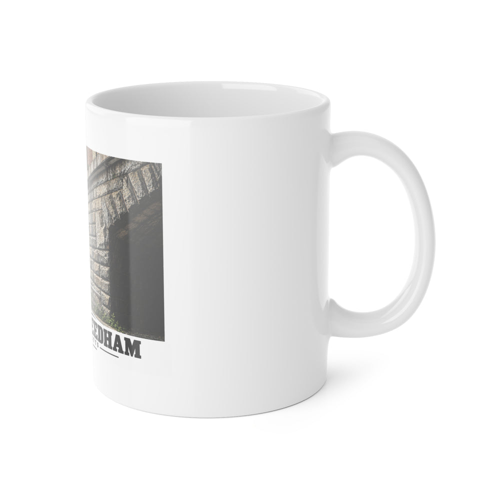 White ceramic mug with its handle on the right and part of the Echo Bridge artwork showing a row of stone arches and a brick upper band, with Newton/Needham lettering visible on the left.