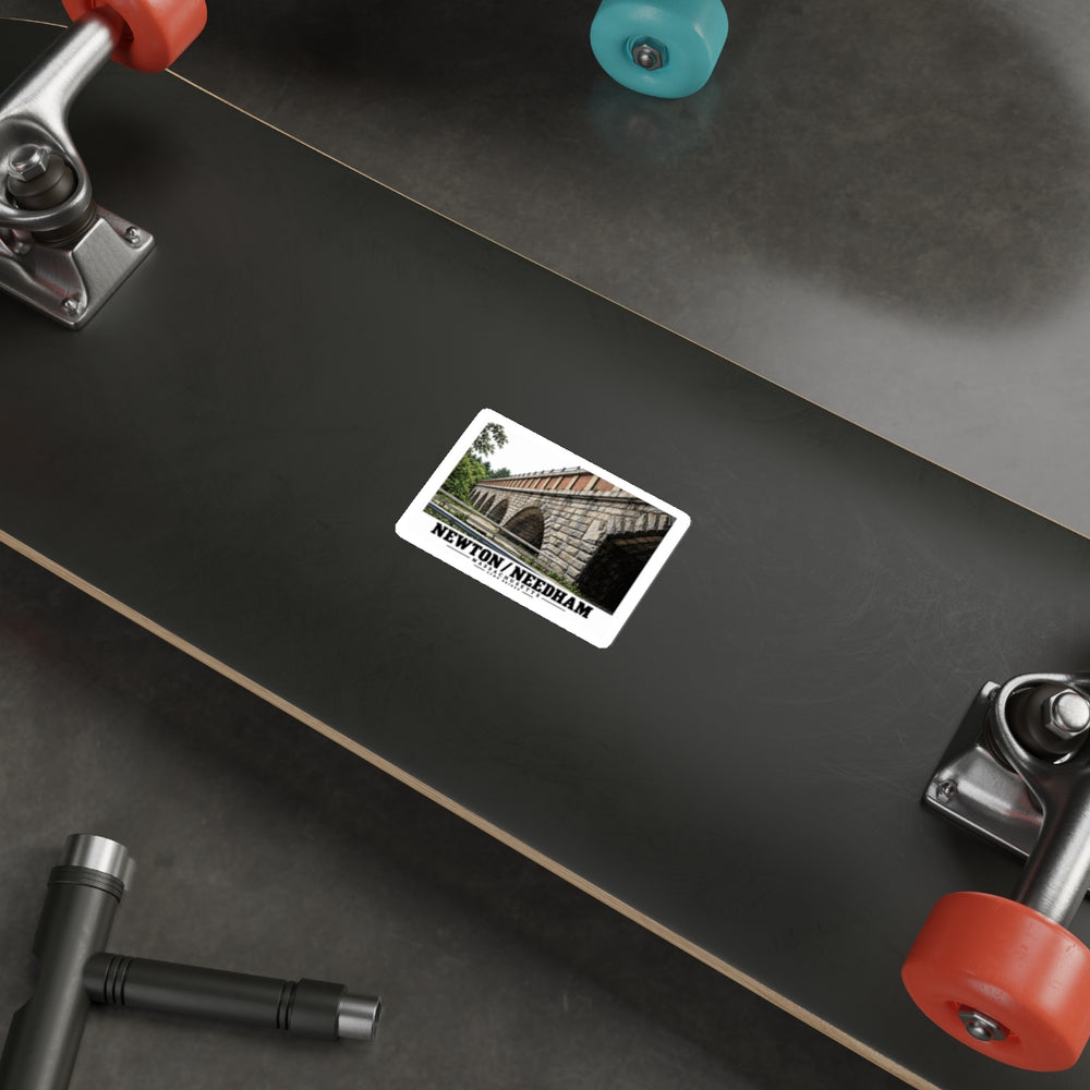 Sticker showing Echo Bridge multi-arch design with Newton/Needham lettering on the underside of a black skateboard with metal trucks and colored wheels