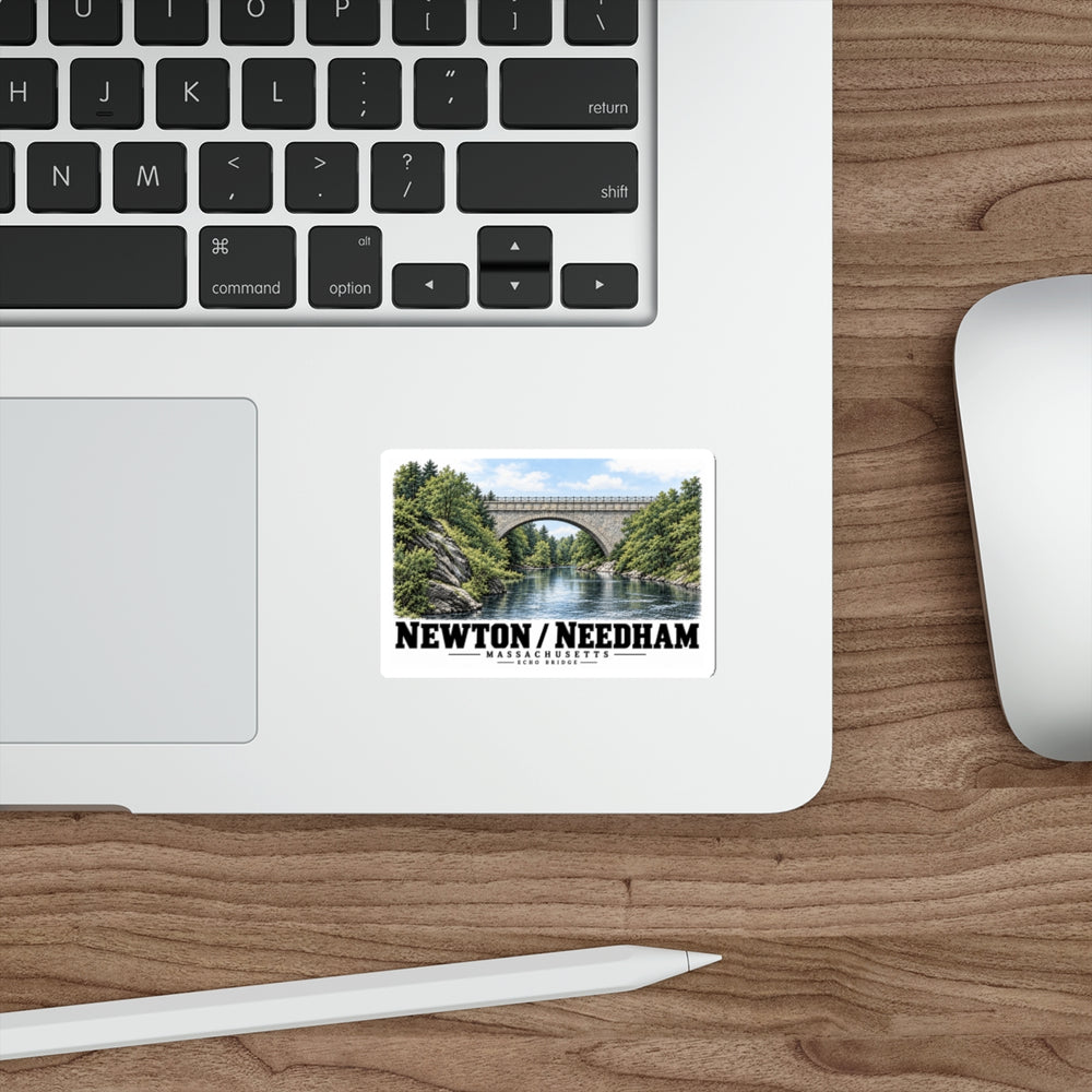 Sticker showing Echo Bridge stone arch and river with Newton/Needham lettering on a laptop palm rest beside the trackpad on a wood desk