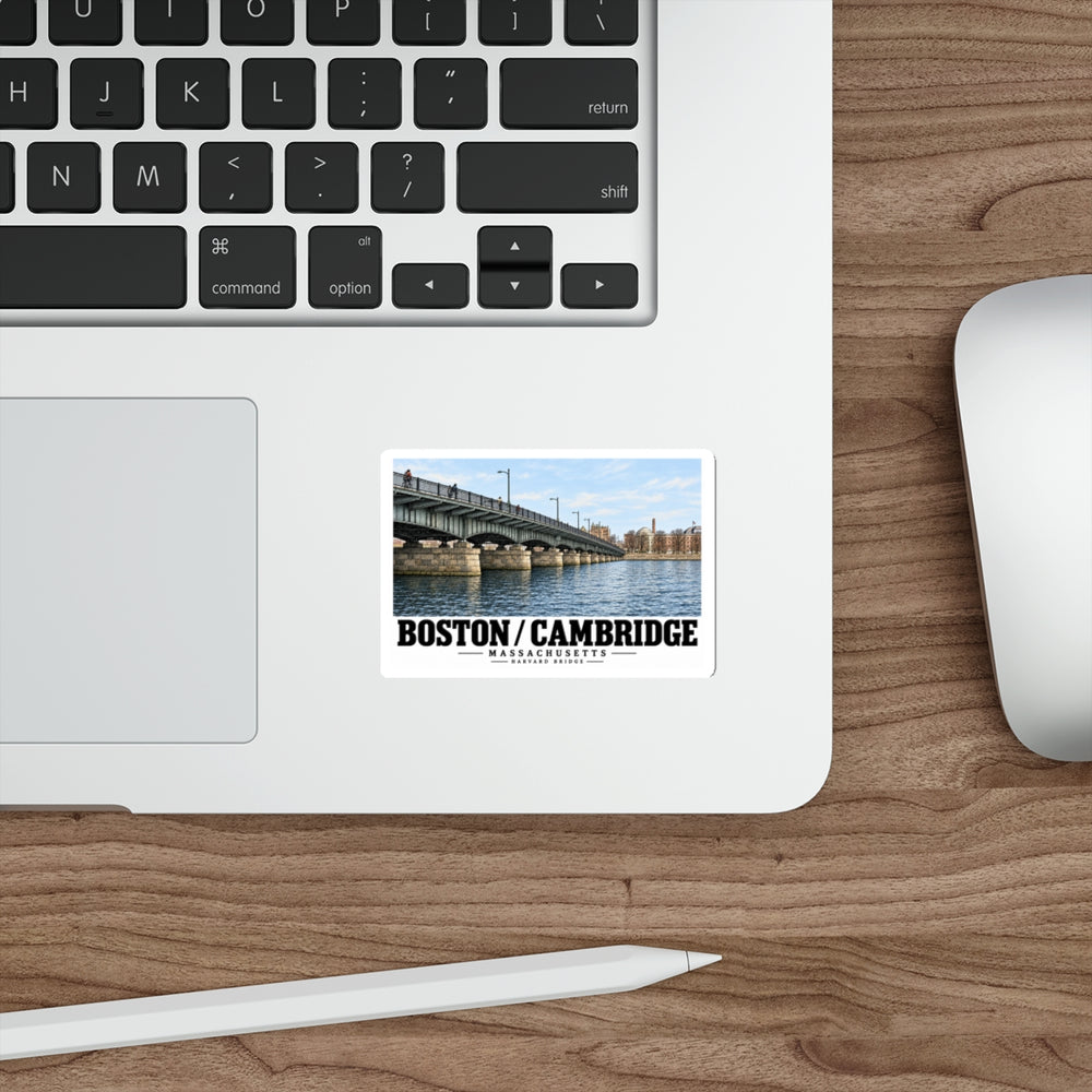 Sticker showing Harvard Bridge spanning the river toward distant buildings with Boston/Cambridge lettering on a laptop palm rest beside the trackpad on a wood desk