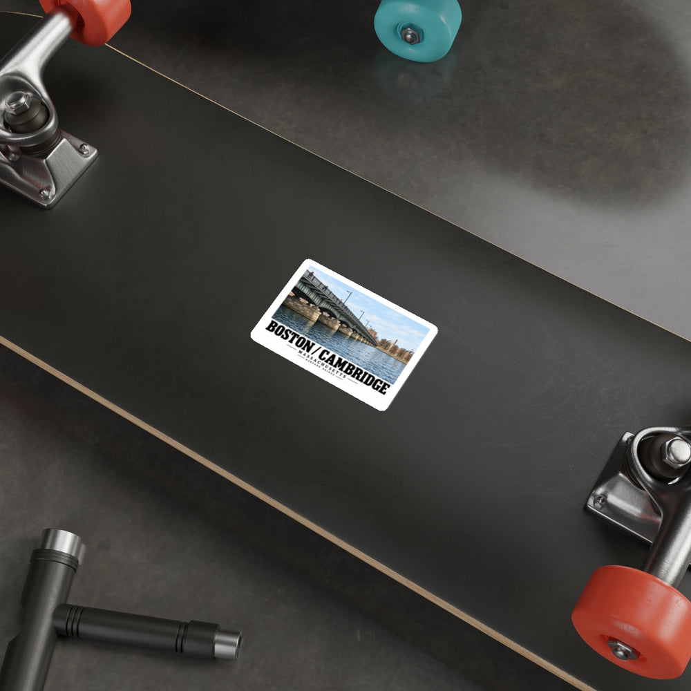 Sticker showing Harvard Bridge spanning the river toward distant buildings with Boston/Cambridge lettering on the underside of a black skateboard with metal trucks and colored wheels