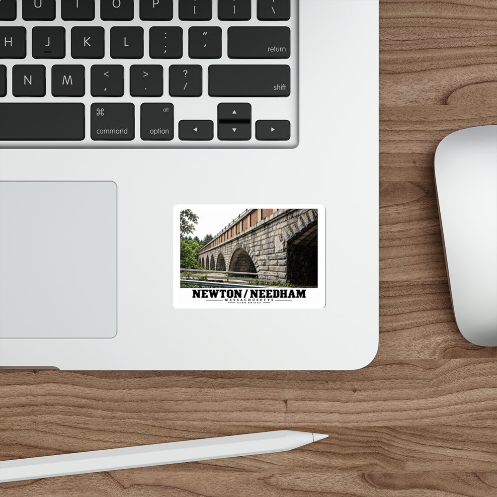 Sticker showing Echo Bridge multi-arch design with Newton/Needham lettering on a laptop palm rest beside the trackpad on a wood desk