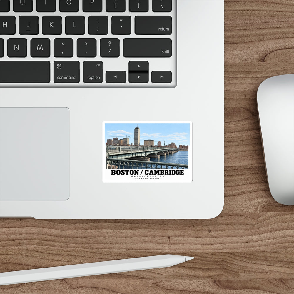 Sticker showing Harvard Bridge and skyline with Boston/Cambridge lettering on a laptop palm rest beside the trackpad on a wood desk