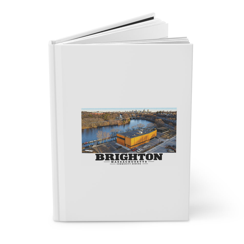 Partly opened white hardcover journal showing Community Rowing aerial building and river artwork on the front